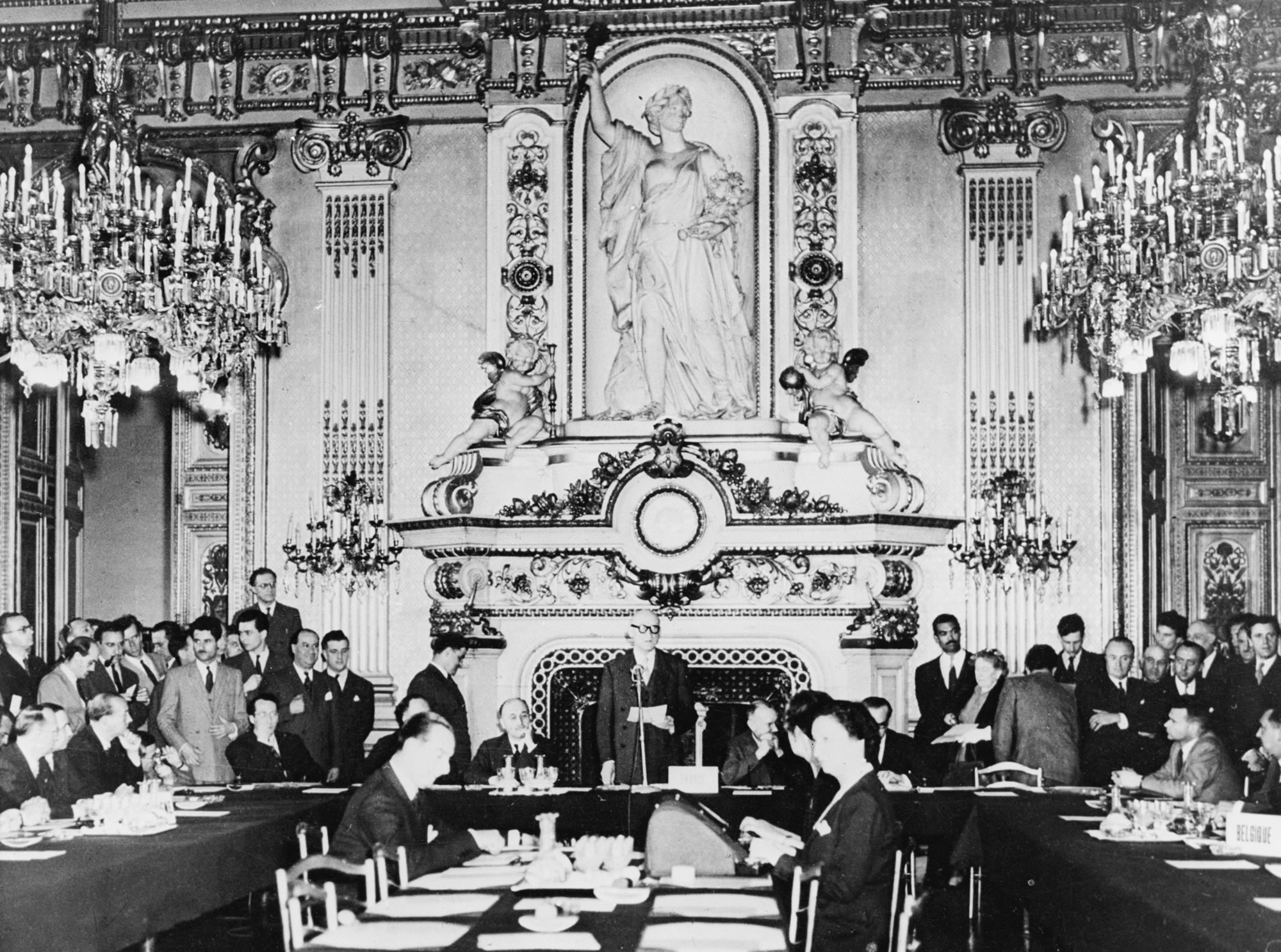 Robert Schuman, é ina sheasamh i lár seomra maorga, os comhair tinteán saothraithe, ag labhairt le toscairí atá ina suí os a chomhair nó ina seasamh timpeall an bhoird.
